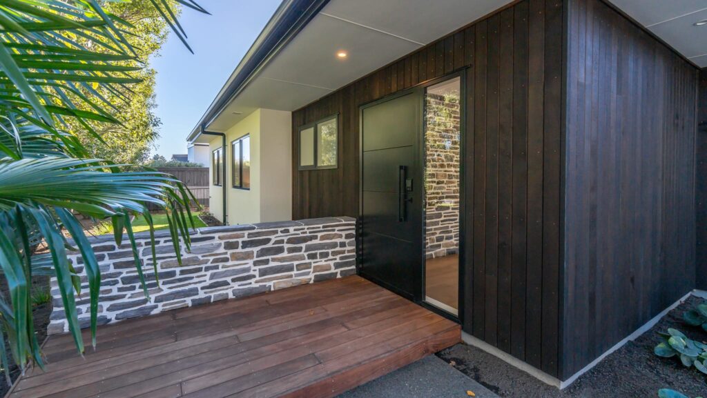 Entrance to a modern house featuring dark timber cladding, stone detailing, and landscaped gardens, ideal for buyers searching for modern houses for sale, architectural homes, new build homes, house and land packages, luxury homes, and property for sale.